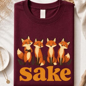 May include: A maroon crew neck t-shirt featuring a graphic of four orange foxes standing in a row above the large, bold, yellow text that reads "For Fox Sake". The foxes have varied expressions and are positioned against a solid dark red background.