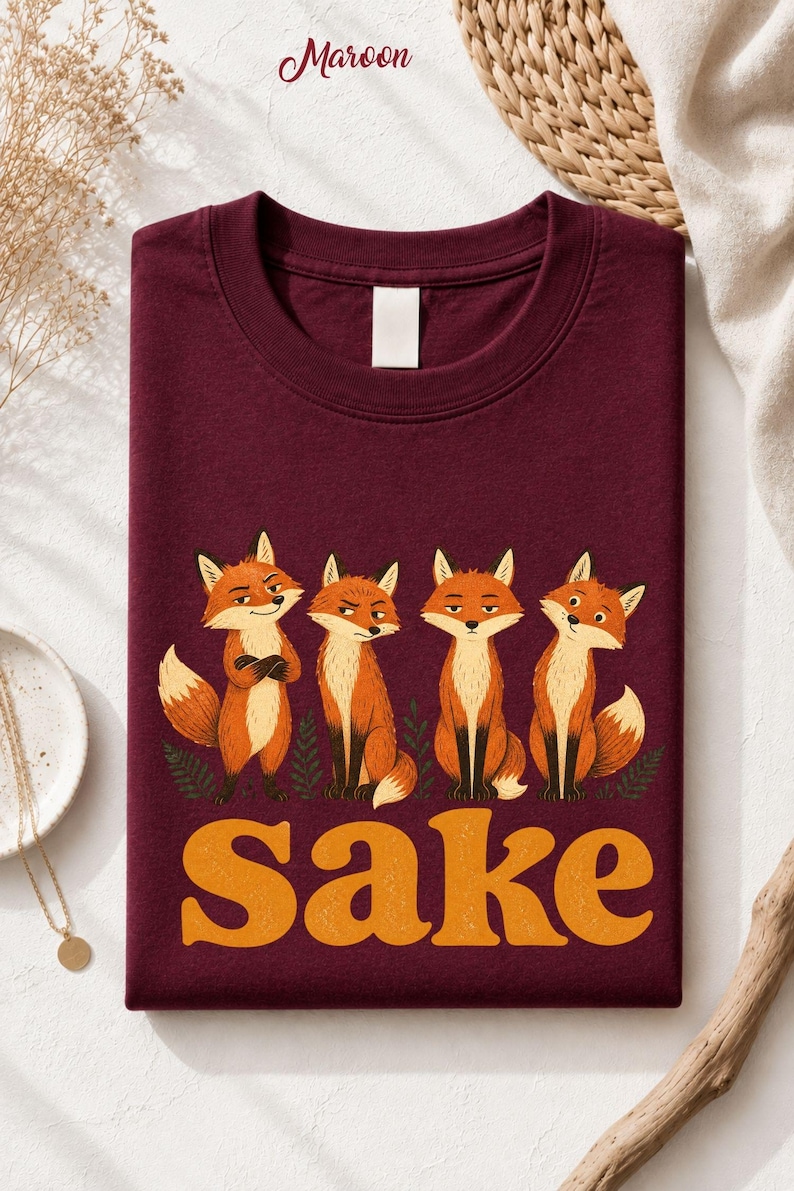 May include: A maroon crew neck t-shirt featuring a graphic of four orange foxes standing in a row above the large, bold, yellow text that reads "For Fox Sake". The foxes have varied expressions and are positioned against a solid dark red background.