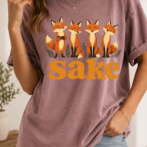 May include: A mauve, short-sleeved, crew-neck t-shirt featuring a graphic of four orange and white foxes standing among green ferns. Below the foxes, the word "sake" is printed in large, bold, orange, retro-style lowercase letters.