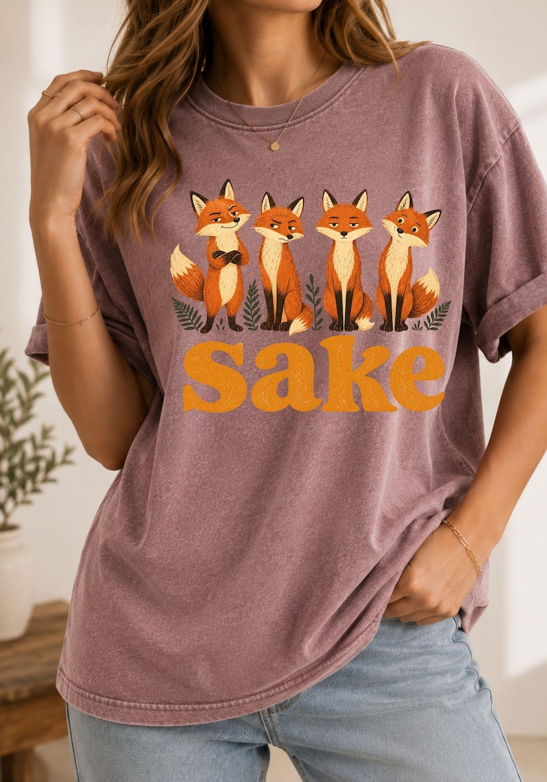 May include: A mauve, short-sleeved, crew-neck t-shirt featuring a graphic of four orange and white foxes standing among green ferns. Below the foxes, the word "sake" is printed in large, bold, orange, retro-style lowercase letters.