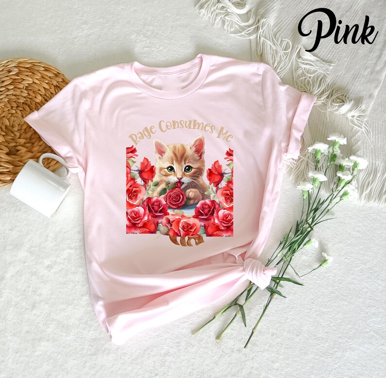 May include: A light pink t-shirt featuring a graphic of a ginger kitten surrounded by red roses. The text "Page Consumes Me" is arched above the kitten. The word "Pink" is written in cursive in the upper right corner.