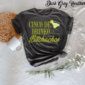 May include: Dark grey heather t-shirt with the text "Cinco De Drinko Bitchachos" in green lettering. A green margarita glass with a lime wedge is next to the text.