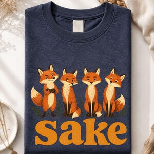 May include: A heather navy crewneck t-shirt featuring a graphic of four orange foxes standing in a row with varying expressions. Below the foxes, the word "Sake" is printed in large, bold, orange, textured, retro-style lettering.