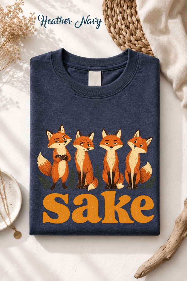 May include: A heather navy crewneck t-shirt featuring a graphic of four orange foxes standing in a row with varying expressions. Below the foxes, the word "Sake" is printed in large, bold, orange, textured, retro-style lettering.