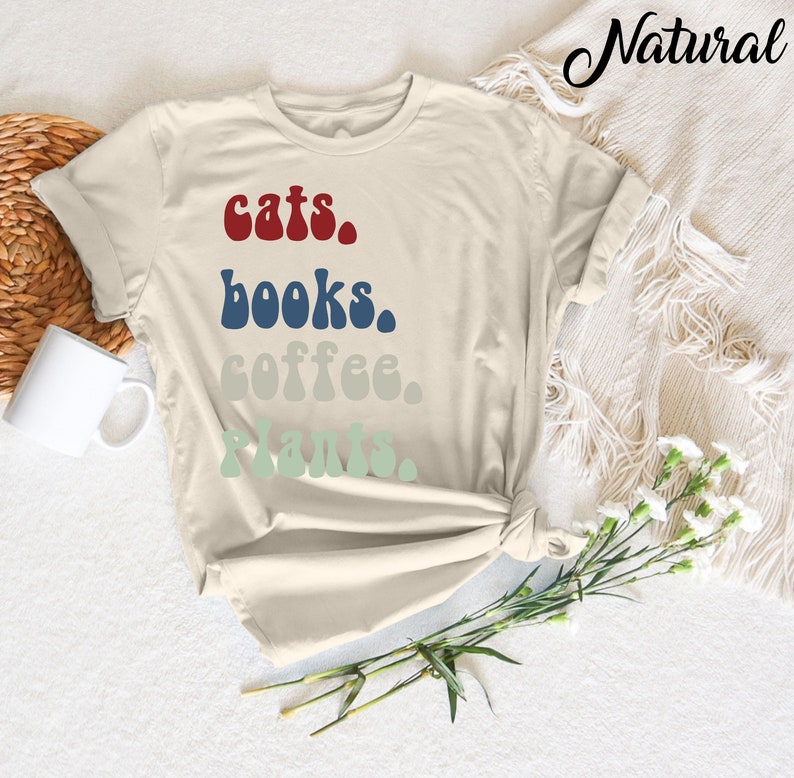 Pu&ograve; includere: Una maglietta beige con il testo "cats. books. coffee. plants." stampato in un font retr&ograve;. Il testo &egrave; in tonalit&agrave; di rosso, blu e verde.