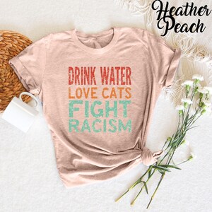 May include: A heather peach colored t-shirt with the text "Drink Water Love Cats Fight Racism" in a vintage style font.