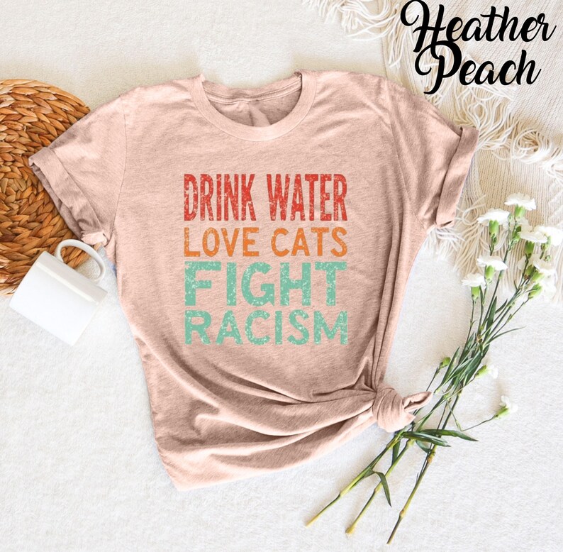 May include: A heather peach colored t-shirt with the text "Drink Water Love Cats Fight Racism" in a vintage style font.