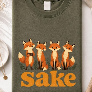 May include: An olive green crew neck t-shirt featuring a graphic of four orange foxes standing in a row above the word "Sake" printed in large, bold, orange letters. The foxes have varied expressions and are surrounded by small green fern illustrations.