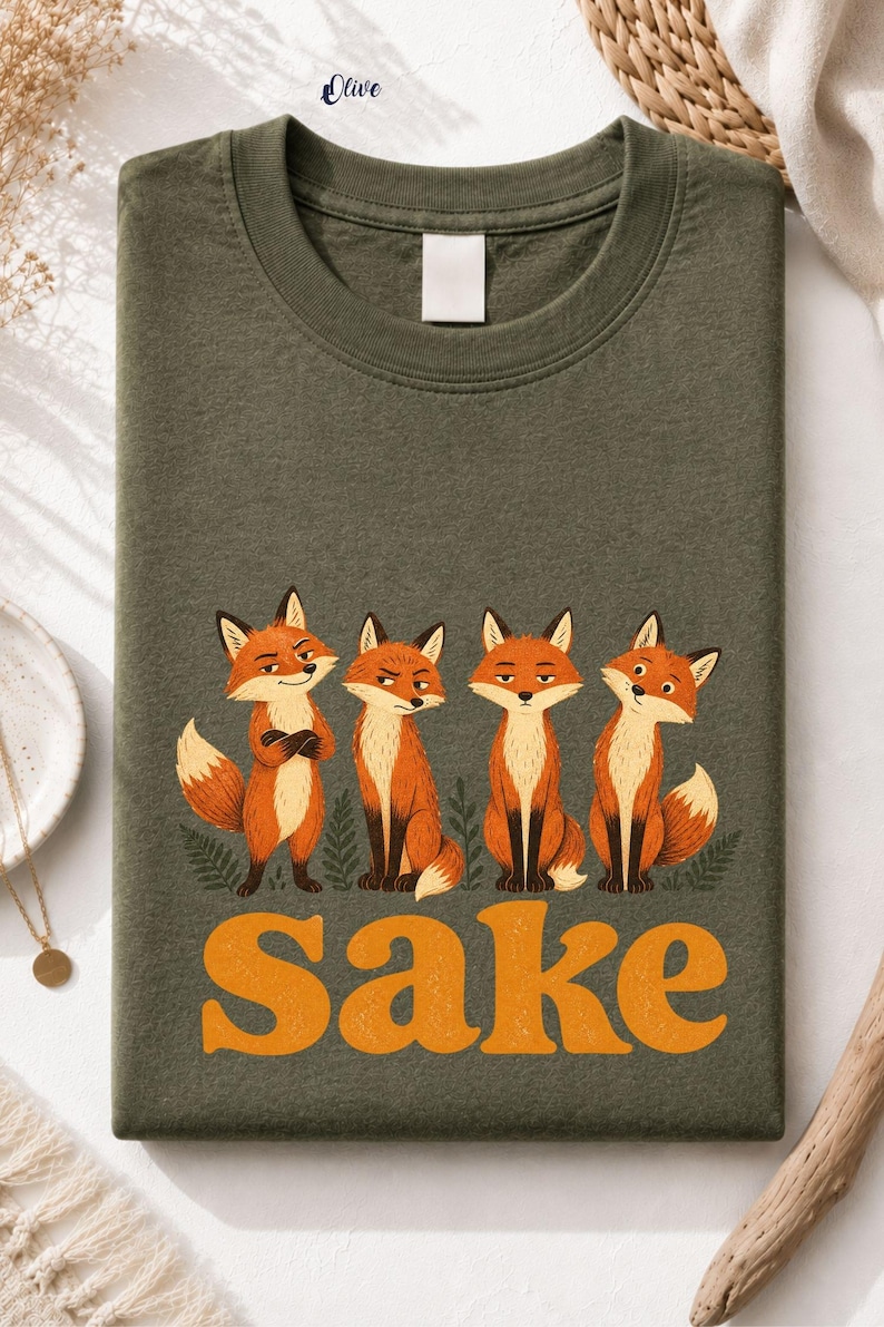 May include: An olive green crew neck t-shirt featuring a graphic of four orange foxes standing in a row above the word "Sake" printed in large, bold, orange letters. The foxes have varied expressions and are surrounded by small green fern illustrations.