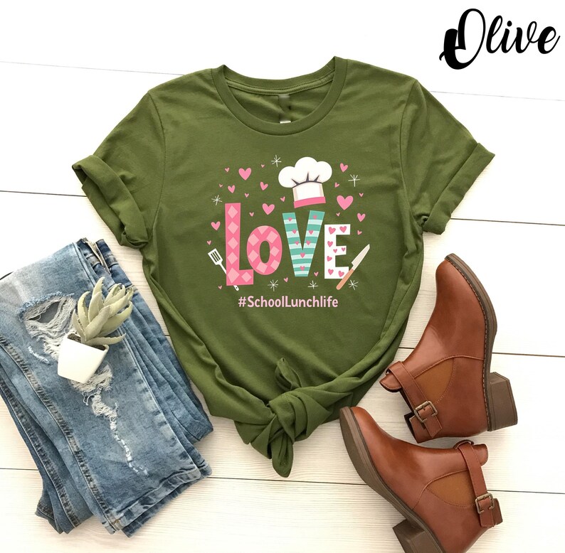 Pode incluir: Camiseta verde oliva com um gr&aacute;fico de um chap&eacute;u de cozinheiro e a palavra "LOVE" em letras rosa e azul. O texto "#SchoolLunchlife" est&aacute; impresso abaixo do gr&aacute;fico.