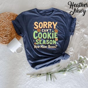 May include: A heather navy blue t-shirt with the text "Sorry can't cookie season. How many boxes?" in green and white lettering. The text is surrounded by cookie images.