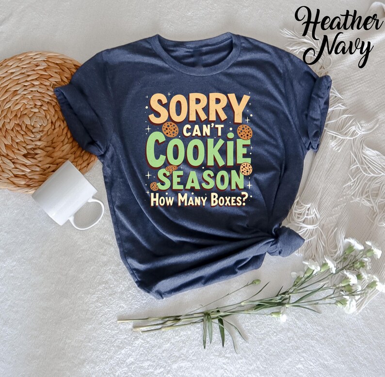 May include: A heather navy blue t-shirt with the text "Sorry can't cookie season. How many boxes?" in green and white lettering. The text is surrounded by cookie images.