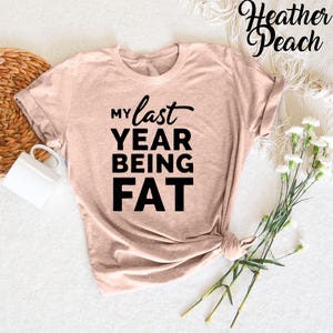 May include: A heather peach colored t-shirt with the text "My last year being fat" printed in black.