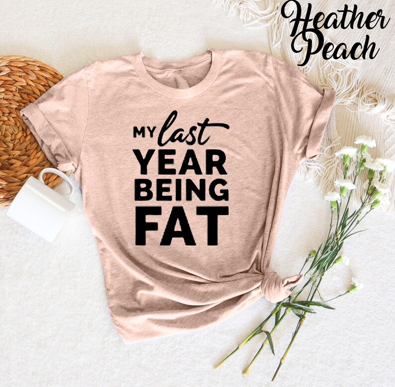 May include: A heather peach colored t-shirt with the text "My last year being fat" printed in black.