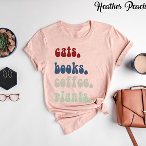 Pu&ograve; includere: Una maglietta rosa con il testo "cats. books. coffee. plants." in un font retr&ograve;. Il testo &egrave; in diversi colori, con "cats" in rosso, "books" in blu, "coffee" in beige e "plants" in verde.