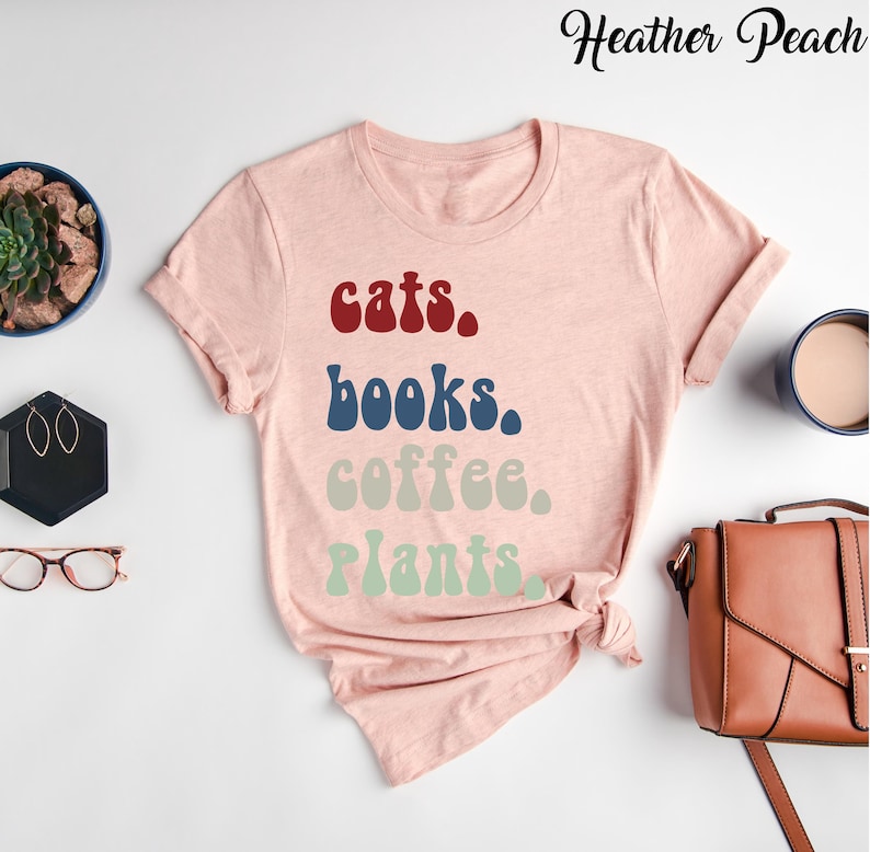 Pu&ograve; includere: Una maglietta rosa con il testo "cats. books. coffee. plants." in un font retr&ograve;. Il testo &egrave; in diversi colori, con "cats" in rosso, "books" in blu, "coffee" in beige e "plants" in verde.