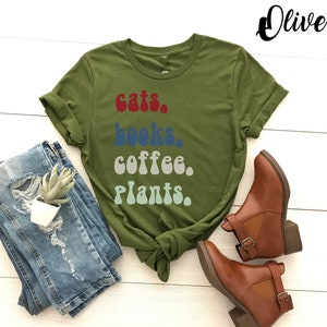 Pu&ograve; includere: Una maglietta verde con il testo "gatti, libri, caff&egrave;, piante." in un font retr&ograve;. Il testo &egrave; in diversi colori, tra cui rosso, blu, grigio e verde.