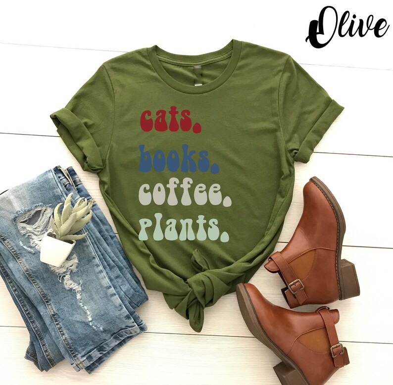 Pu&ograve; includere: Una maglietta verde con il testo "gatti, libri, caff&egrave;, piante." in un font retr&ograve;. Il testo &egrave; in diversi colori, tra cui rosso, blu, grigio e verde.