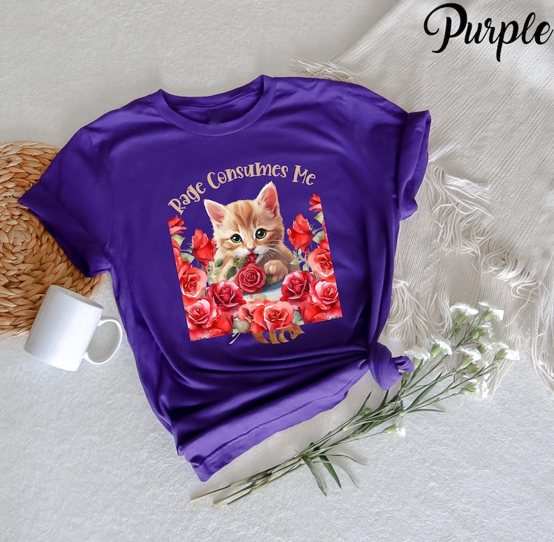 May include: A purple t-shirt featuring a graphic of a ginger kitten surrounded by red roses. The text "Rage Consumes Me" is arched above the kitten. The word "Purple" is written in cursive in the upper right corner.