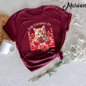 May include: Maroon t-shirt featuring a graphic of a kitten surrounded by red roses, with the text "Rage Consumes Me". The shirt is made of a soft material and has short sleeves. A white mug and decorative flowers are in the background.