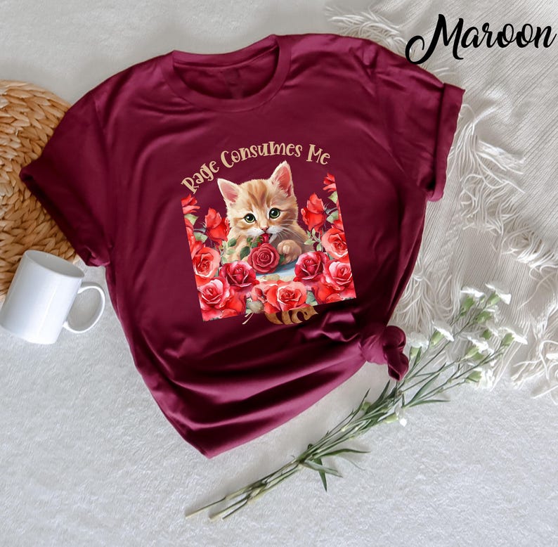 May include: Maroon t-shirt featuring a graphic of a kitten surrounded by red roses, with the text "Rage Consumes Me". The shirt is made of a soft material and has short sleeves. A white mug and decorative flowers are in the background.