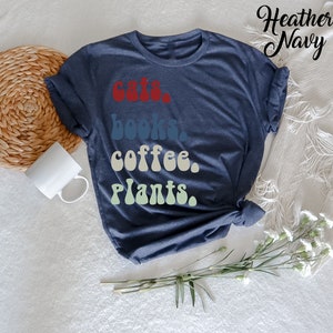 Pu&ograve; includere: Una t-shirt blu navy heather con il testo "gatti, libri, caff&egrave;, piante." stampato in un font retr&ograve; con un effetto sfumato.