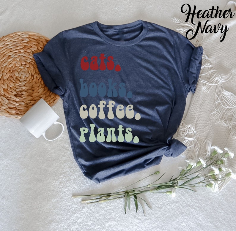 Pu&ograve; includere: Una t-shirt blu navy heather con il testo "gatti, libri, caff&egrave;, piante." stampato in un font retr&ograve; con un effetto sfumato.
