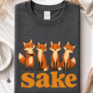 May include: A dark heather gray crew neck t-shirt featuring a graphic of four orange foxes with various expressions. Below the foxes, the word "Sake" is printed in large, bold, orange, textured letters. The shirt has a plain white neck label.