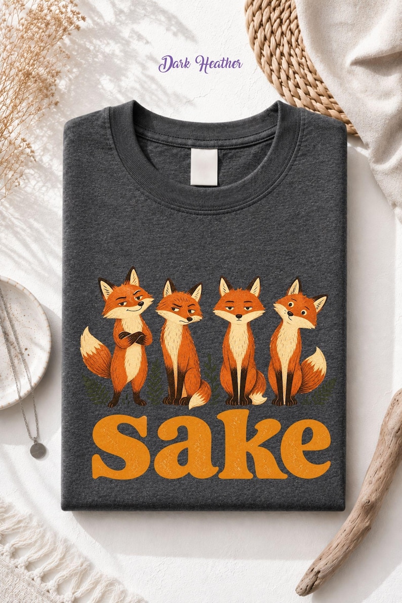 May include: A dark heather gray crew neck t-shirt featuring a graphic of four orange foxes with various expressions. Below the foxes, the word "Sake" is printed in large, bold, orange, textured letters. The shirt has a plain white neck label.