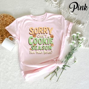 May include: Pink t-shirt with the text "Sorry can't cookie season. How many boxes?" in green and yellow letters. The text is surrounded by images of chocolate chip cookies.