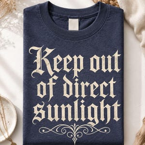 Op de afbeelding: Een heather navy t-shirt met de tekst "Keep out of direct sunlight" in een cr&egrave;mekleurig gotisch lettertype. Onder de tekst staat een decoratieve krul. Het t-shirt heeft een ronde hals en een leeg label bij de binnenkant van de kraag.