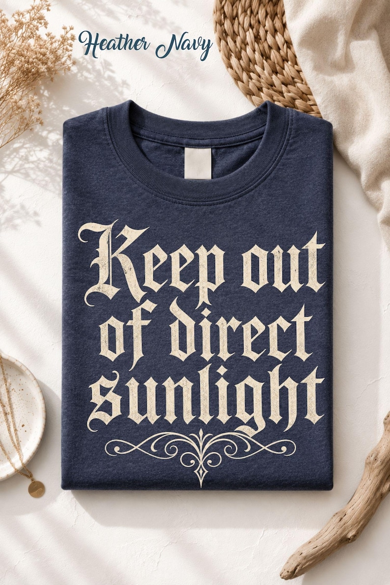 Op de afbeelding: Een heather navy t-shirt met de tekst "Keep out of direct sunlight" in een cr&egrave;mekleurig gotisch lettertype. Onder de tekst staat een decoratieve krul. Het t-shirt heeft een ronde hals en een leeg label bij de binnenkant van de kraag.