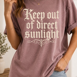 Op de afbeelding: Een stoffig mauvekleurig t-shirt met korte mouwen en de tekst "Keep out of direct sunlight" in een cr&egrave;mekleurig schreeflettertype op de borst, vergezeld van een decoratief krulontwerp onder de tekst.