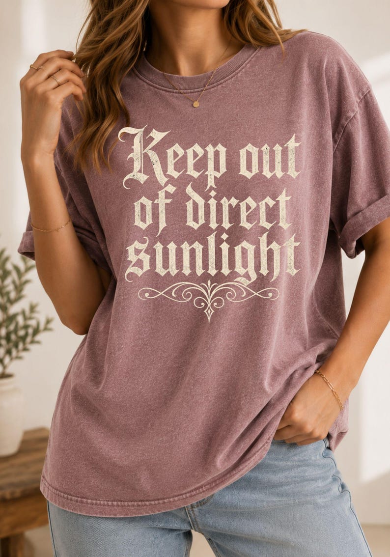 Op de afbeelding: Een stoffig mauvekleurig t-shirt met korte mouwen en de tekst "Keep out of direct sunlight" in een cr&egrave;mekleurig schreeflettertype op de borst, vergezeld van een decoratief krulontwerp onder de tekst.
