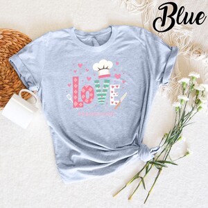 Pode incluir: Uma camiseta azul claro com um design gr&aacute;fico que diz "LOVE" em letras coloridas com um chap&eacute;u de cozinheiro e utens&iacute;lios de cozinha. O texto abaixo do gr&aacute;fico diz "#school lunchlife".