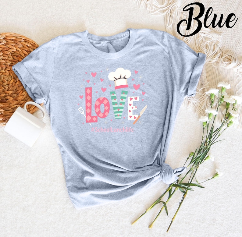 Pode incluir: Uma camiseta azul claro com um design gr&aacute;fico que diz "LOVE" em letras coloridas com um chap&eacute;u de cozinheiro e utens&iacute;lios de cozinha. O texto abaixo do gr&aacute;fico diz "#school lunchlife".