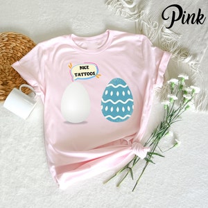 May include: A light pink t-shirt featuring two Easter egg designs. One egg is white, and the other is blue with a wavy pattern. The text "NICE TATTOOS" is in a speech bubble above the eggs. The word "Pink" is written in cursive.
