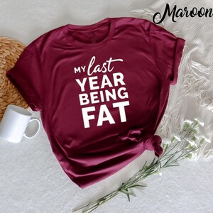 May include: A maroon t-shirt with the text "My last year being fat" in white.