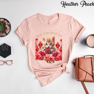 May include: A light pink t-shirt featuring a watercolor illustration of a ginger kitten surrounded by red roses. The text "Page Consumes Me" is arched above the kitten. The words "Heather Peach" are written in cursive at the top.