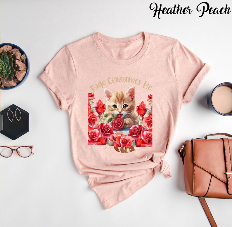 May include: A light pink t-shirt featuring a watercolor illustration of a ginger kitten surrounded by red roses. The text "Page Consumes Me" is arched above the kitten. The words "Heather Peach" are written in cursive at the top.