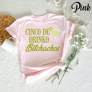 May include: Pink t-shirt with the text "Cinco de Drinko Bitchachos" in green lettering and a green margarita glass with a lime wedge.
