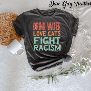 May include: Dark grey heather t-shirt with the text "Drink Water Love Cats Fight Racism" in a vintage style font.