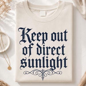 Op de afbeelding: Een natuurkleurig t-shirt met de donkerblauwe tekst Keep out of direct sunlight in een gotisch lettertype. Het ontwerp bevat een decoratieve krul onder de tekst. Het t-shirt heeft een ronde hals en een leeg wit label bij de kraag.