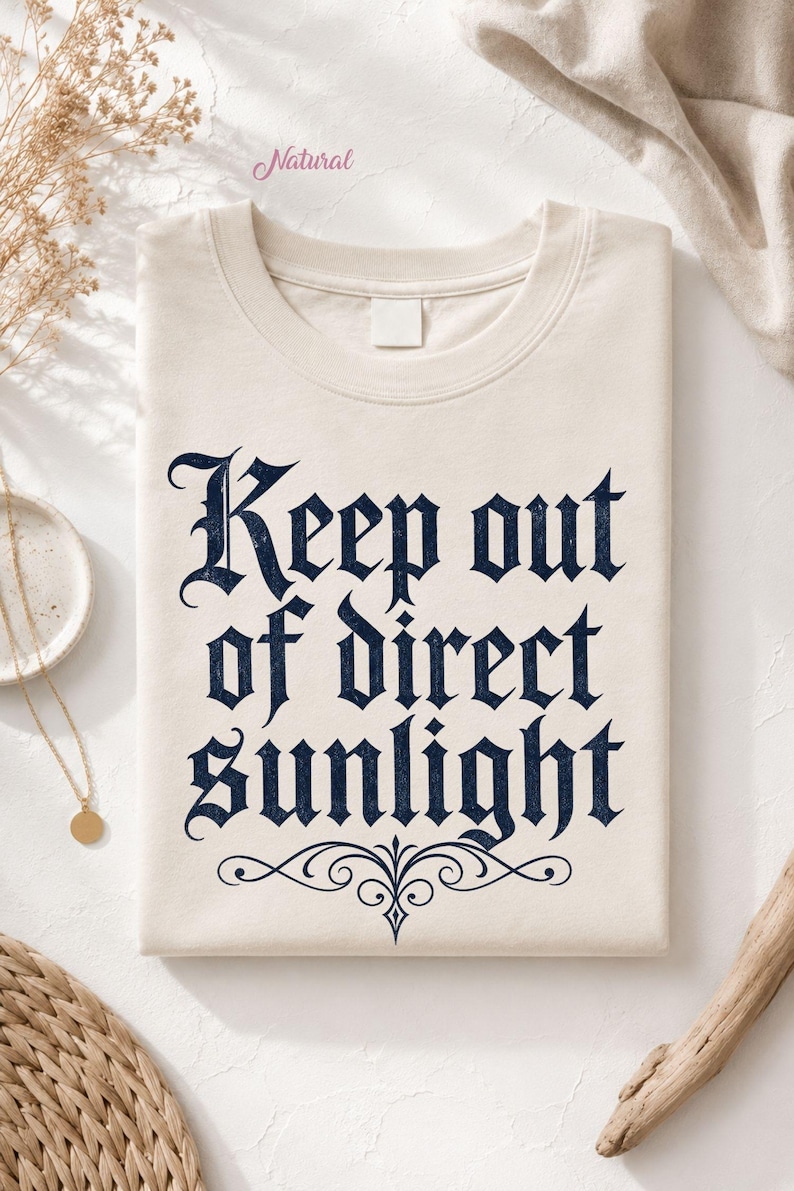 Op de afbeelding: Een natuurkleurig t-shirt met de donkerblauwe tekst Keep out of direct sunlight in een gotisch lettertype. Het ontwerp bevat een decoratieve krul onder de tekst. Het t-shirt heeft een ronde hals en een leeg wit label bij de kraag.