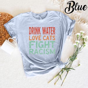 May include: A light blue t-shirt with the text "Drink Water Love Cats Fight Racism" in a vintage style font. The text is in red, orange, and green.