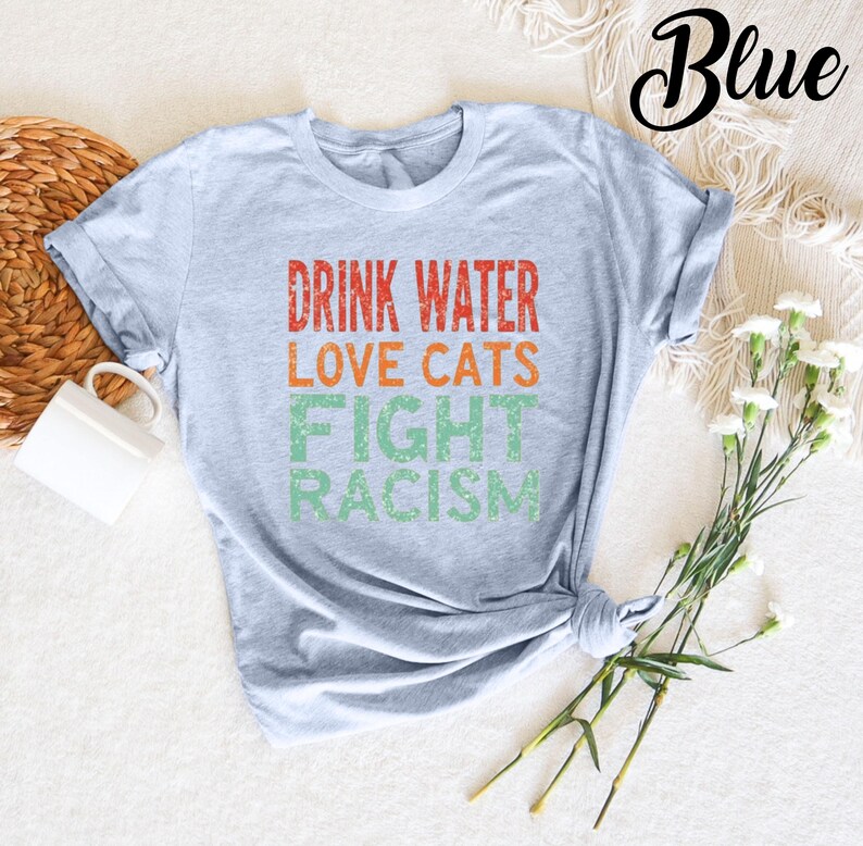 May include: A light blue t-shirt with the text "Drink Water Love Cats Fight Racism" in a vintage style font. The text is in red, orange, and green.
