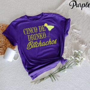 May include: Purple t-shirt with the text "Cinco de Drinko Bitchachos" in gold lettering. A green margarita glass with a lime wedge is pictured above the text.