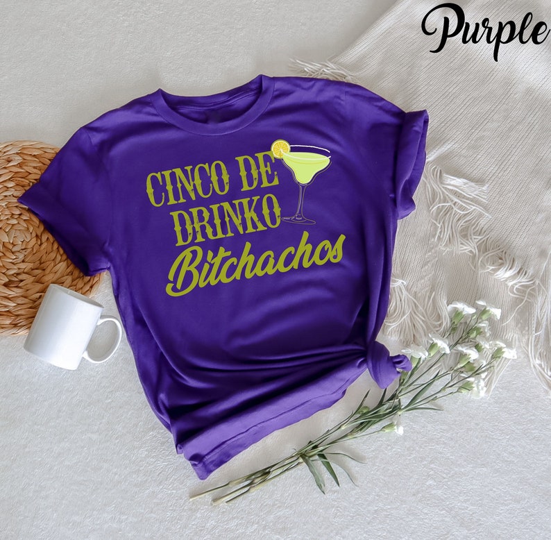 May include: Purple t-shirt with the text "Cinco de Drinko Bitchachos" in gold lettering. A green margarita glass with a lime wedge is pictured above the text.