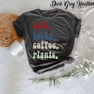 Pu&ograve; includere: Una maglietta in grigio melange scuro con il testo "gatti, libri, caff&egrave;, piante" in un font retr&ograve;. Il testo &egrave; in diversi colori: rosso, blu, beige e verde.