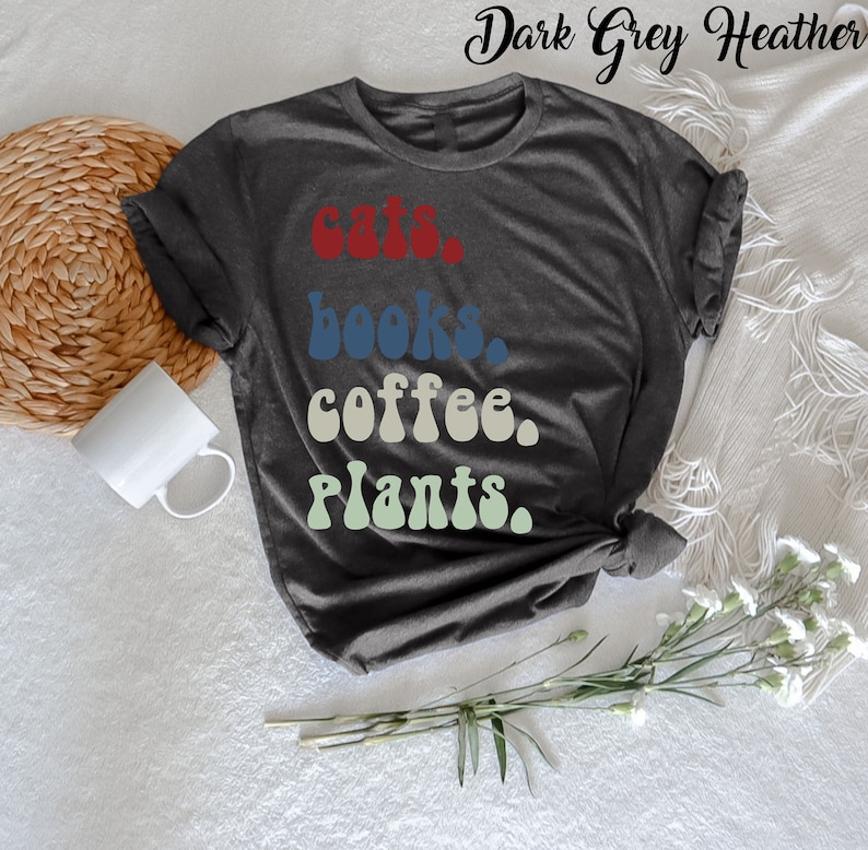 Pu&ograve; includere: Una maglietta in grigio melange scuro con il testo "gatti, libri, caff&egrave;, piante" in un font retr&ograve;. Il testo &egrave; in diversi colori: rosso, blu, beige e verde.